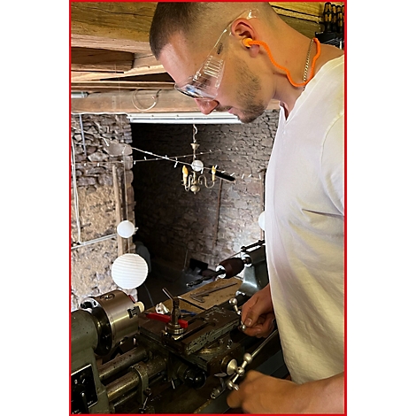 Mann mit Schutzbrille arbeitet an einer Drehmaschine, trägt Gehörschutz. Im Hintergrund: Steinmauer und Kronleuchter.